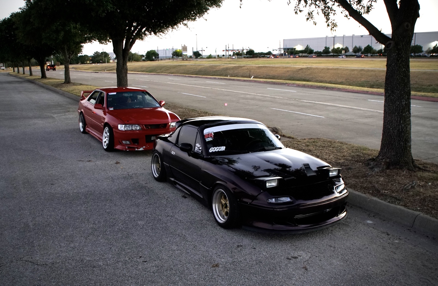 Black Mazda Miata and red Nissan Skyline at a roadside meet